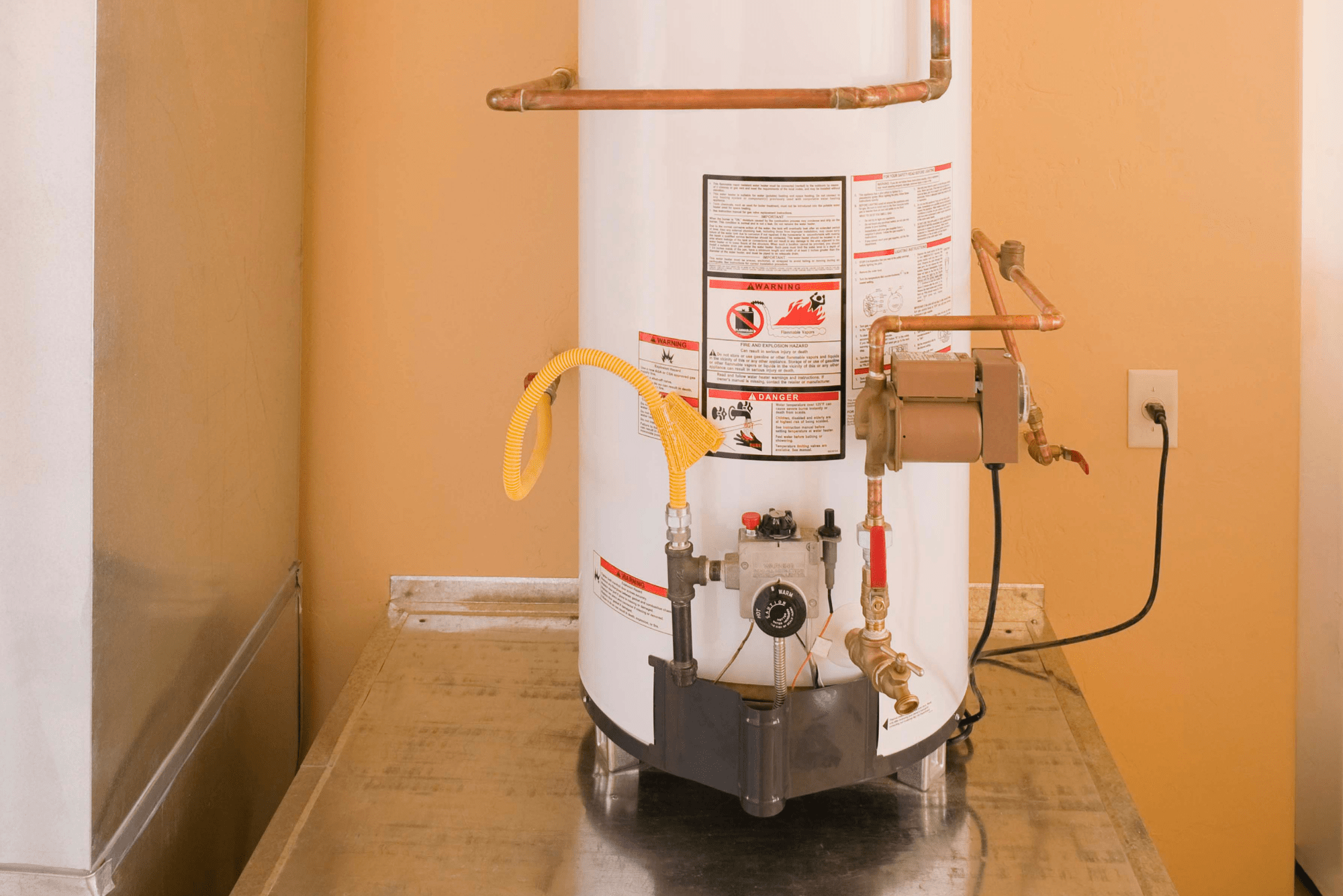 A water heater with copper pipes and a yellow hose, positioned against a light-colored wall, on a metal surface.